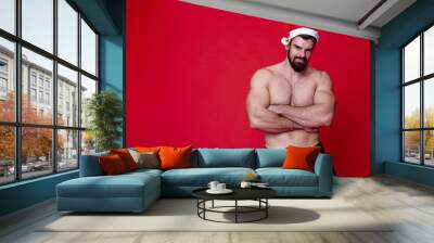 Sexy santa, young shirtless bodybuilder wearing a christmas hat, playing santa claus for holidays, red background studio portrait Wall mural