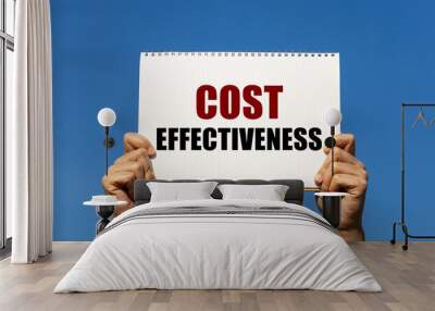 Cost effectiveness text on notebook paper held by 2 hands with isolated blue sky background. This message can be used as business concept about cost effectiveness. Wall mural