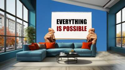 Everything is possible text on notebook paper held by 2 hands with isolated blue sky background. This message can be used as business concept about everything is possible. Wall mural