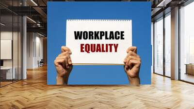 Workplace equality text on notebook paper held by 2 hands with isolated blue sky background. This message can be used as business concept about workplace equality. Wall mural