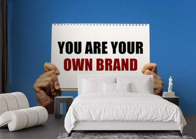 You are your own brand text on notebook paper held by 2 hands with isolated blue sky background. This message can be used as business concept about branding yourself. Wall mural