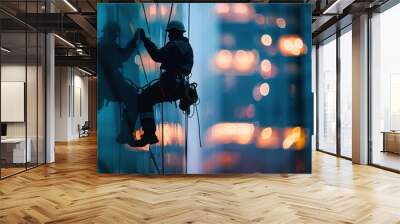 Silhouette of a worker in safety gear performing high-rise window cleaning on a glass building facade at twilight, with city lights in the background. Wall mural