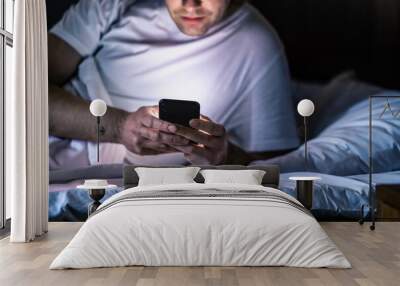 Man in bed with phone at night. Texting with smartphone before sleeping. Guy holding cellphone in hand. Dark home bedroom. Infidelity, cheating or working late online concept. Screen light on face. Wall mural