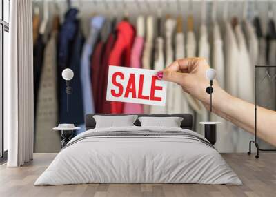 Sale in clothing shop. Clothes hanging in hanger in a rack and wardrobe in a fashion store with discount and bargain prices. Woman holding advertising and marketing banner or discount coupon in hand. Wall mural