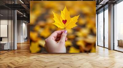 Holding a vibrant yellow maple leaf against a backdrop of fallen autumn foliage in a serene park setting Wall mural