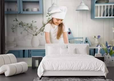 little cute smiling girl wearing a white chef's hat and uniform with tongue kneading the dough for cake in the blue and white kitchen with easter decoration Wall mural