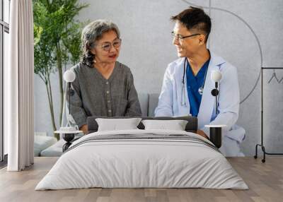 Asian young man doctor holding digital tablet consulting old patient, Male doctor therapist in uniform sit on sofa at hospital or clinic with senior patient during medical visit doing health checkup Wall mural