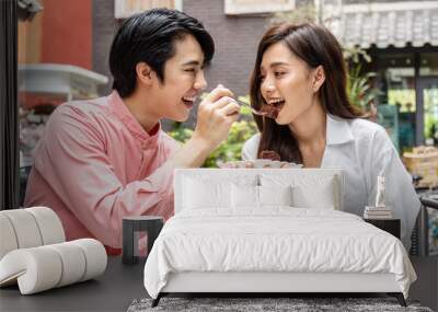 Young asian man feeding happy girlfriend with tasty chocolate cake sitting at table with coffee at date in restaurant Wall mural