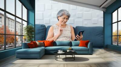 Happy Asian senior woman sit on couch, doing victory sign by hand at digital tablet screen in hand in white room. Elderly female meet with family by video call at home. Older people with technology. Wall mural