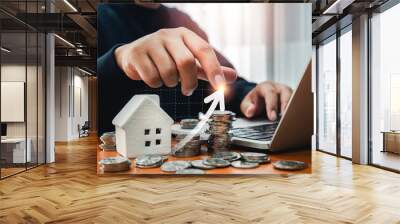 Home investment, savings, income, real estate price value, property profit concepts. Arrow up on graph pointing by man hand while work with laptop computer near white house and coin piles on table. Wall mural