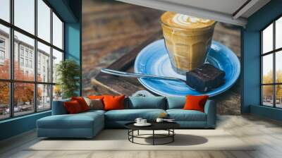 Piccolo Latte coffee topping with flower art from milk in small glass with a piece of homemade brownies cake on blue ceramic plate on wooden table Wall mural