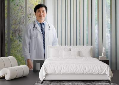 Smiling Asian man doctor portrait standing near the glass windows in medical office with green view. Confident Asian adult male doctor with stethoscope holding blue document folder, looking at camera. Wall mural