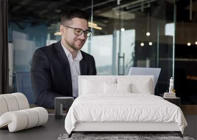 A smiling young man businessman in a business suit sits and works in the office on a laptop online, texts, chats with partners, clients Wall mural