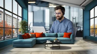 A smiling young man in a blue shirt is sitting at a desk in a modern office, wearing a headset, working and studying at a laptop Wall mural