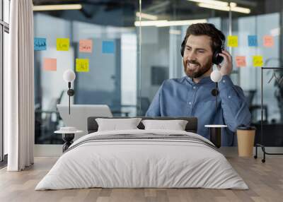 A smiling young man in a headset is working in the office on a laptop, sitting at the workplace and talking remotely through a video call Wall mural