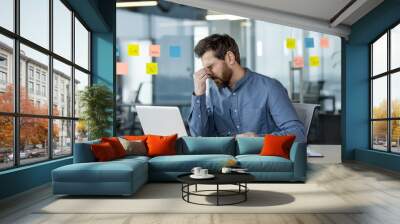 A tired young man in a blue shirt is sitting at the desk in the office and working overtime at the laptop, rubbing his eyes with his hand, holding glasses Wall mural