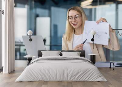 A young business woman works in the office at a laptop, talks on a video call with the command, partners, director. Presents and shows the camera documents with graphs, financial income Wall mural