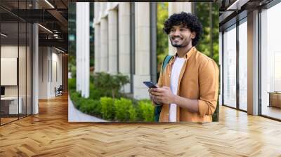 A young Indian male programmer stands outside on the street with a backpack and uses the phone. He looks away with a smile Wall mural