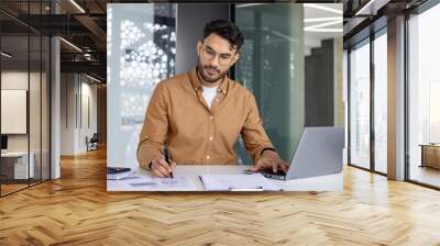 A young Muslim man is working concentratedly in the office at the table with a laptop, making notes of data and documents Wall mural