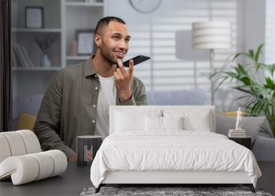 African american young man sitting on sofa at home and talking on speaker phone, recording voice message, dictating his conversation on mobile app Wall mural