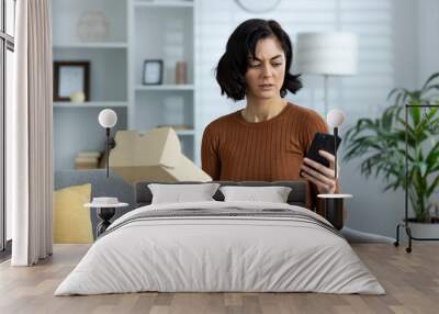 Angry and upset young woman is at home, sitting on the sofa, holding an open parcel box and calling on a mobile phone to the delivery service and customer service center with a complaint Wall mural