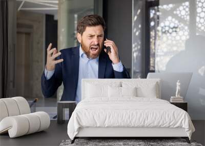 Angry young male businessman sitting at a desk in the office and talking aggressively on the phone, shouting and gesturing with his hands Wall mural