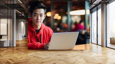 Asian young man in red hoodie working on laptop, freelancer boy or student with computer in cafe at table, looking at camera. IT specialist works remotely using laptop while sitting in cafe Wall mural