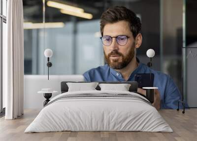 Close-up photo of a serious and focused man in glasses and a shirt working in the office on a laptop and holding a mobile phone Wall mural