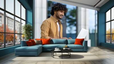 Close-up photo of a smiling young Indian man using a laptop while sitting on a street bench Wall mural