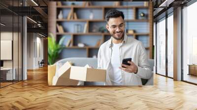 Close-up photo of an Indian smiling young man who received a parcel, holds a box in his hands and dials on a mobile phone. Sitting on the couch at home Wall mural