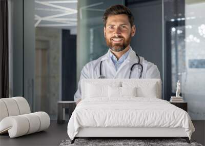 Close-up portrait of a smiling young male doctor, assistant, student standing in the hospital office and confidently looking at the camera with his arms crossed on his chest Wall mural