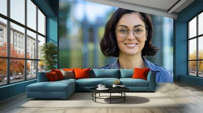 Close-up portrait of a young beautiful woman in glasses and a shirt, a student smiling and looking at the camera outside a university campus on a sunny day Wall mural