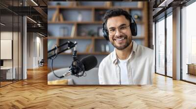 Close-up portrait of a young Indian man wearing glasses and headphones at home, sitting in front of a microphone and looking at the camera Wall mural