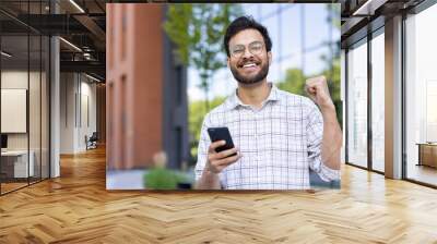 Close-up portrait of a young Muslim man standing outside a building, holding a phone, making a hand gesture and smiling at the camera Wall mural