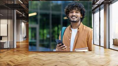 Close-up portrait of young smiling Muslim male student holding books and mobile phone and looking at camera Wall mural