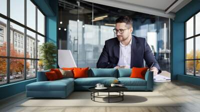 Concentrated young businessman office worker working at workplace with documents. Looks at the laptop monitor and takes notes Wall mural