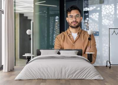 Confident young man with glasses standing and smiling in a modern office environment Wall mural