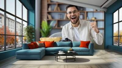 Excited man celebrating video game win at home while holding game controller and smiling on couch Wall mural