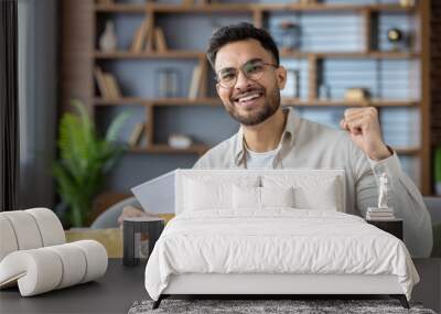 Excited young man celebrating receipt of good news in letter, with happy expression and fist pump, sitting in cozy modern home interior Wall mural