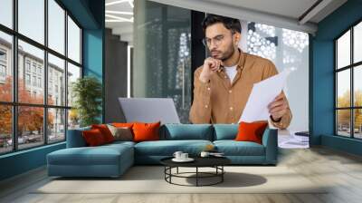 Focused businessman working on laptop and holding documents in modern office Wall mural