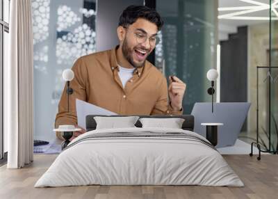 Happy Indian man sitting in office at desk, holding documents in hand, looking at laptop screen and happy with achievement and result Wall mural