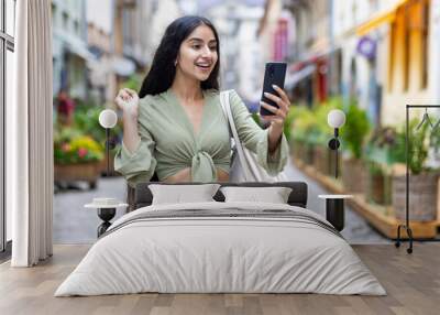 Happy Indian young woman standing on city street in green suit and looking at phone screen, happy to receive news and message showing victory gesture with hand Wall mural