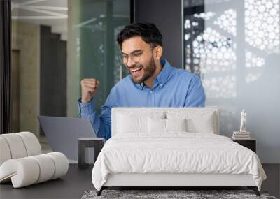 Happy young Indian man working in the office, sitting with a chair and enjoying success, looking at the laptop screen and showing a victory gesture with his hand Wall mural