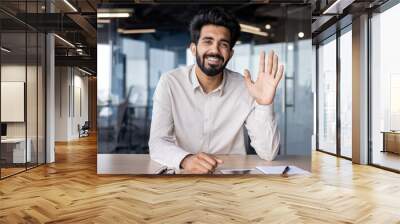 Online business meeting and training. Indian young businessman sitting at the desk in the office, waving to the camera and smiling Wall mural