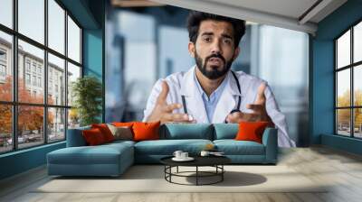 Online medical consultation. Portrait of a serious young Indian male doctor sitting in the office at the table talking, consulting the camera, gesturing with his hands Wall mural