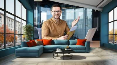 Portrait of a man sitting in the office and working on a laptop. Received and holds in his hands an envelope with a letter and documents. Smiling looking at the camera Wall mural