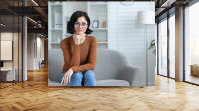 Portrait of a serious young woman in glasses and casual clothes sitting on the sofa at home, looking at the camera with concentration and seriousness. holds his hand near his chin Wall mural