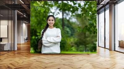 Portrait of a smiling Indian young girl in a white sweater doing sports and walking in the park. He stands with his hands crossed on his chest and looks confidently into the camera Wall mural