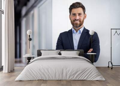 Portrait of a smiling young successful male businessman standing in the office in a suit and looking at the camera with his arms crossed on his chest Wall mural