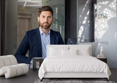 Portrait of a successful and confident young man lawyer in a business suit sitting at an office desk, working with a laptop and documents, looking seriously at the camera Wall mural